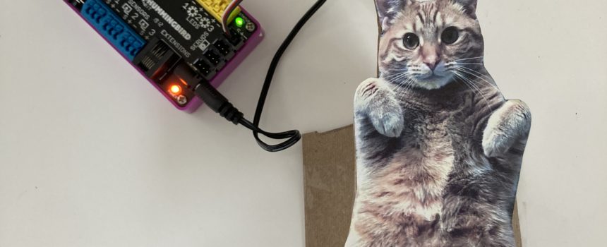 A Hummingbird Bit sits, connected to the power pack, with the micro:bit displaying a happy face on its LED array. A servo motor is plugged into the Bit controller. The Bit is connected to a cutout of a plump orange tabby cat.